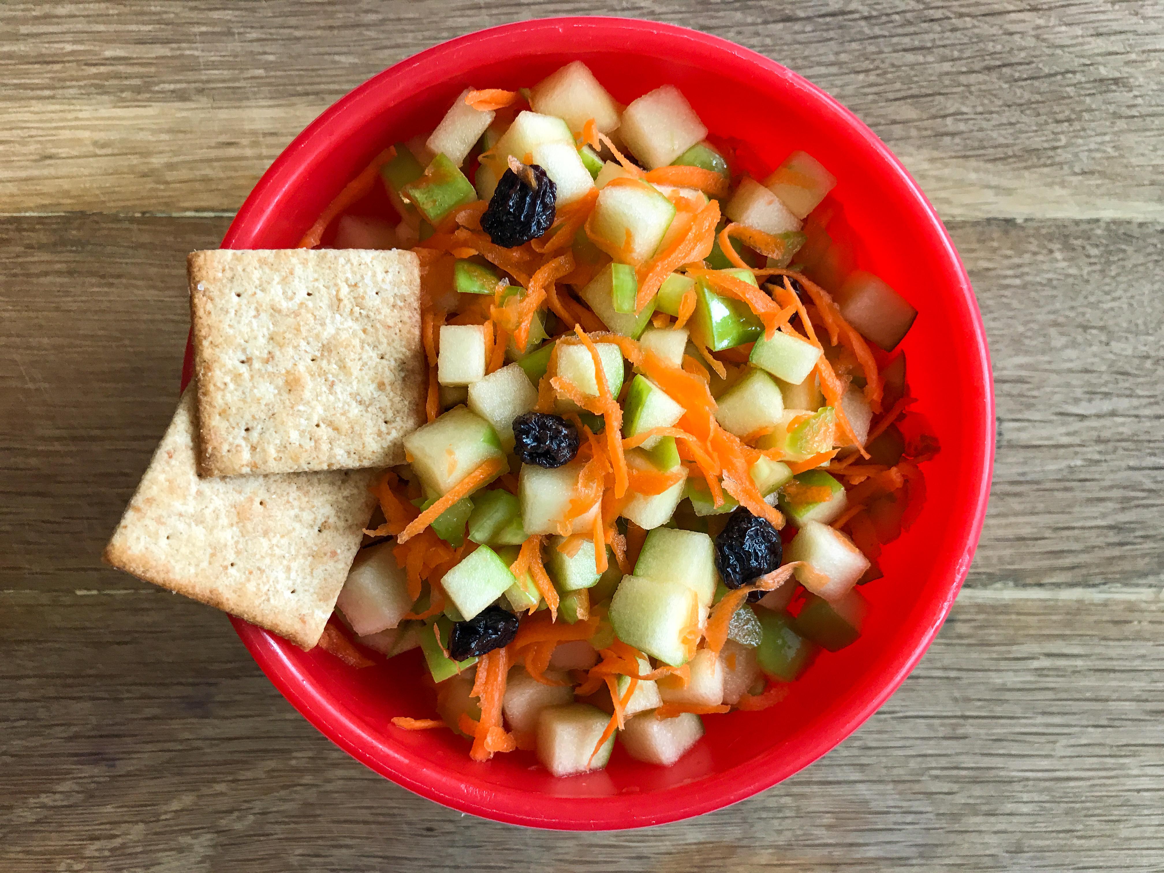 apple cubes topped with raisins and carrot shreds in a bowl with 2 crackers
