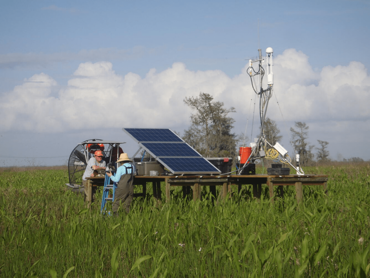 A tall metal eddy covariance tower standing in a field, equipped with sensors at the top to measure wind, temperature, humidity, and gas exchange. The tower is surrounded by a growing wheat crop under an open sky, representing its use in measuring evapotranspiration and carbon fluxes between land and atmosphere. Two scientists are standing nearby and appear to be in a conversation.