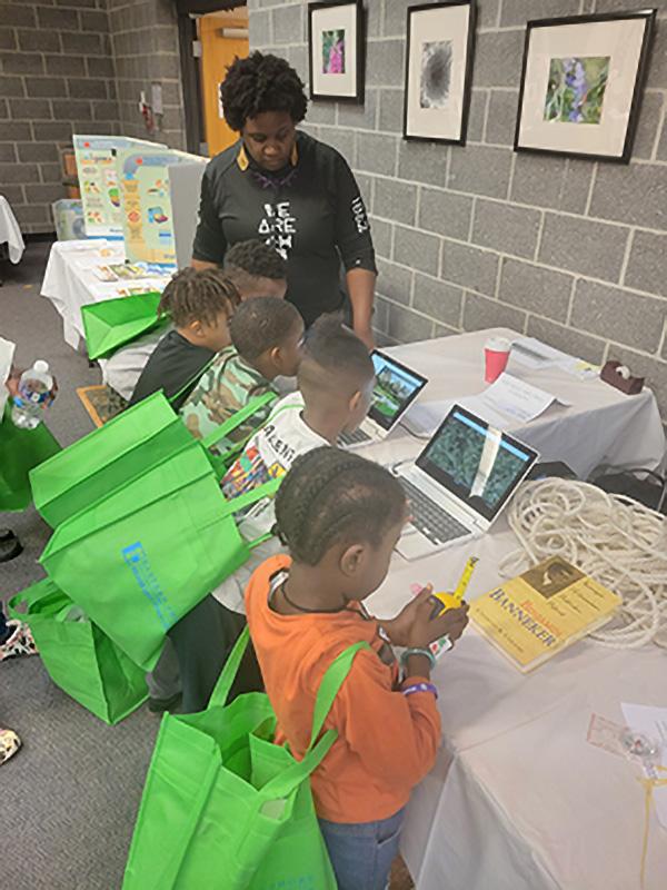  A group of children with green tote bags gather around a table with laptops. An adult guides them.