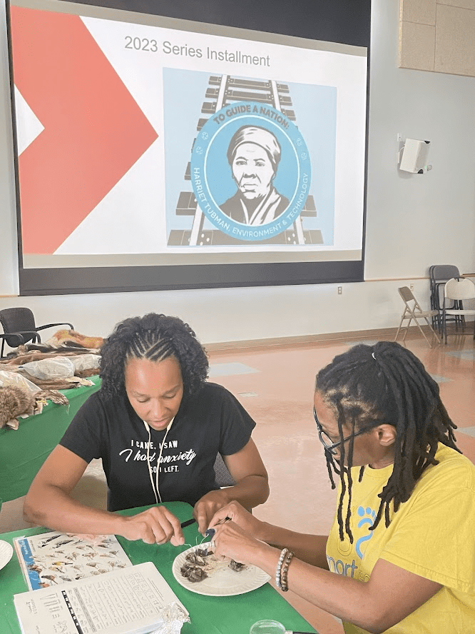 Two people work on a project at a table, with charts and materials. Behind them, a screen displays "2023 Series Installment" with a portrait logo.
