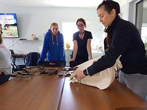 A woman in a black jacket shows a horse skull to three people at a table with horse tack. They appear attentive and focused in a classroom setting.