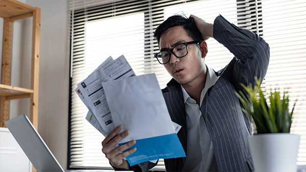 A man in a suit looks confused and stressed while holding multiple bills in an office with blinds and a laptop. A plant is on the desk nearby.