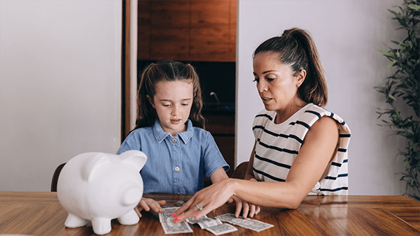A white piggy bank sits on a wooden table in the foreground. Two people, one wearing a blue button-up shirt and the other wearing a striped sleeveless top, are seated at the table. They are engaged in a financial activity, with several dollar bills spread out on the table between them.