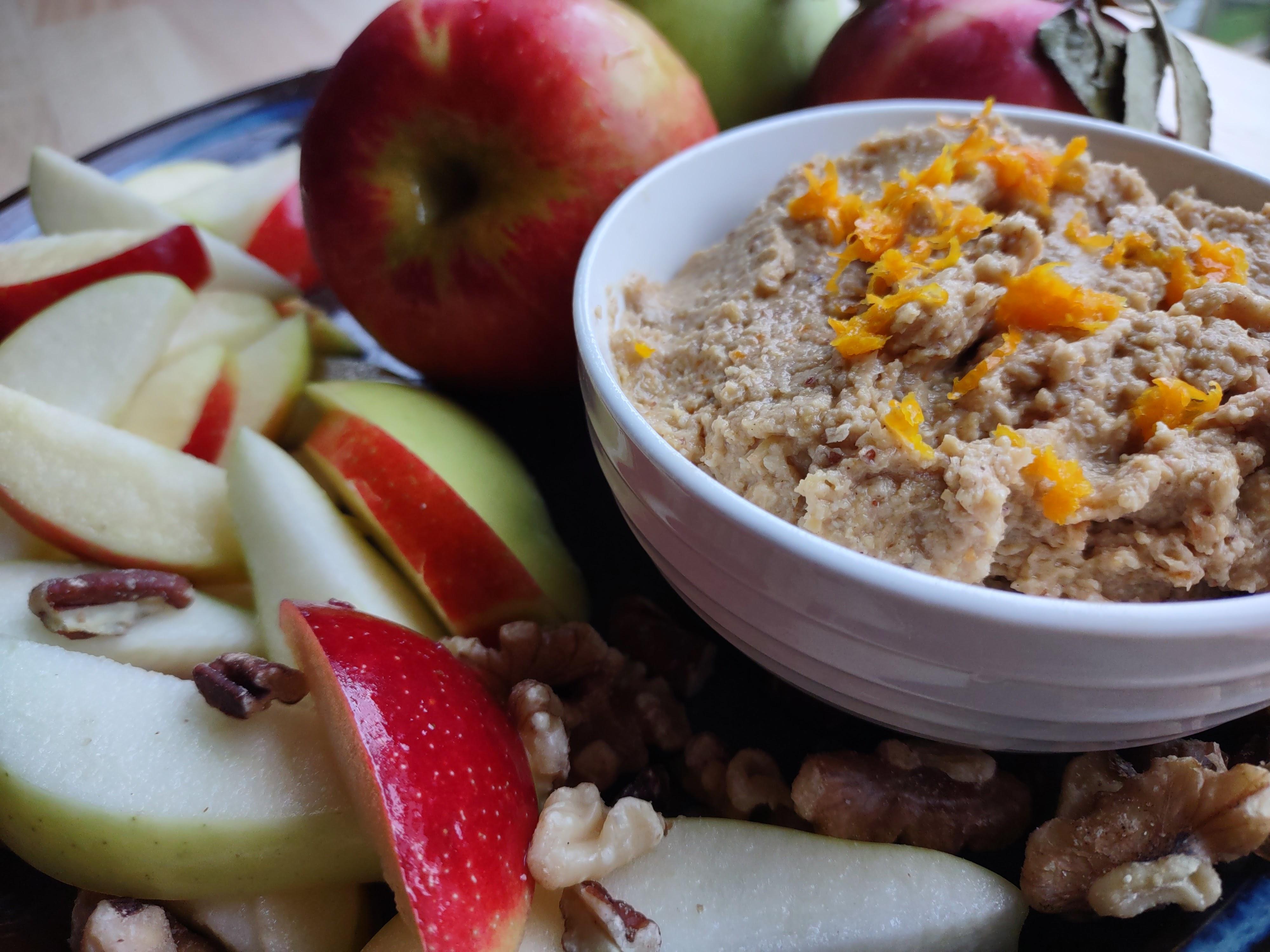 Walnut hummus dip in a white bowl with sliced apples to the left hand side of bowl.