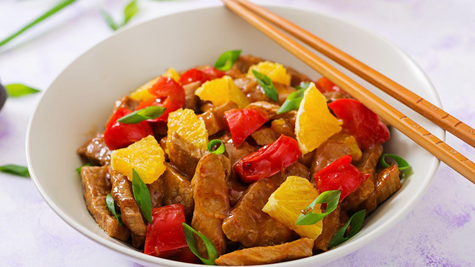 A beef stir fry with red bell peppers and canned oranges . Chopsticks to the right hand side of the bowl.