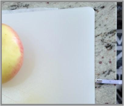 Cutting board with a damp paper towel peaking out from under it. A ruler shows that the cutting board is 1" to 1.5" away from the edge of the counter. 