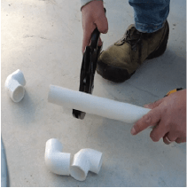 A person is cutting one PVC pipe and three PVC elbows scattered on the floor, indicating preparation for the construction of the bell siphon drain.